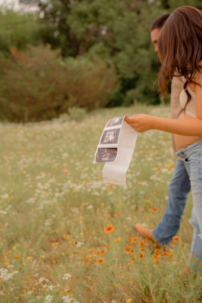 Couple walking across flower field holding baby sonogram