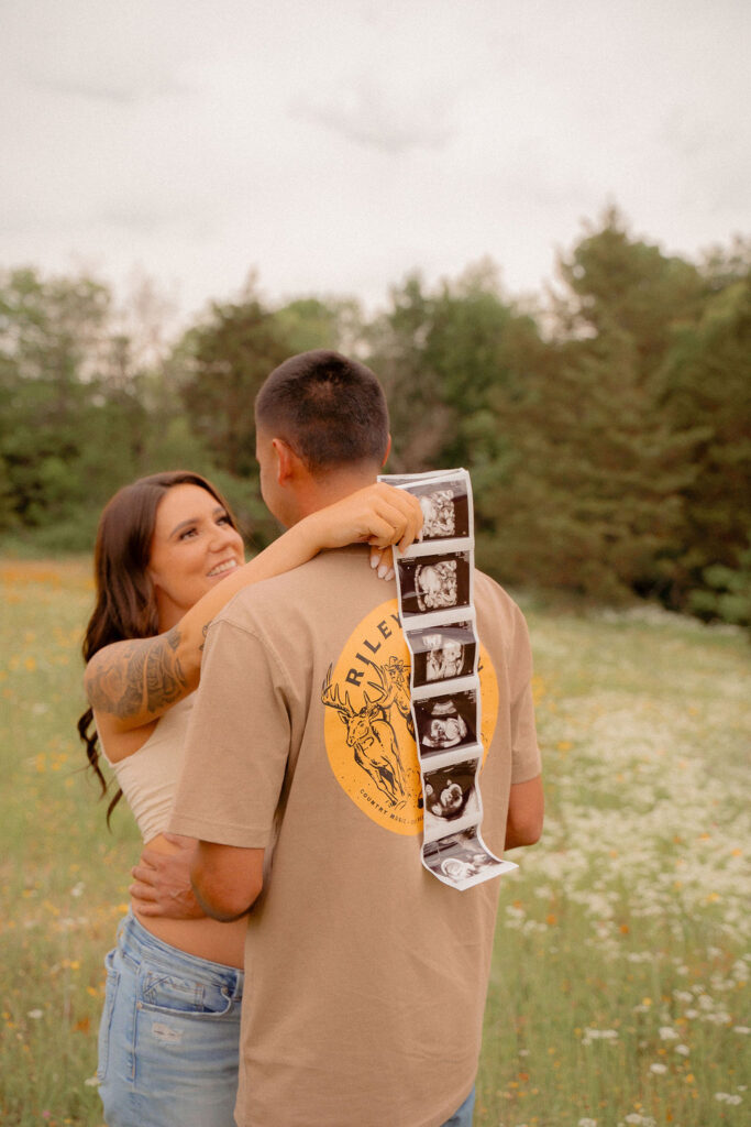 Mom and dad hugging staring in love while holding their sonogram in a flower field