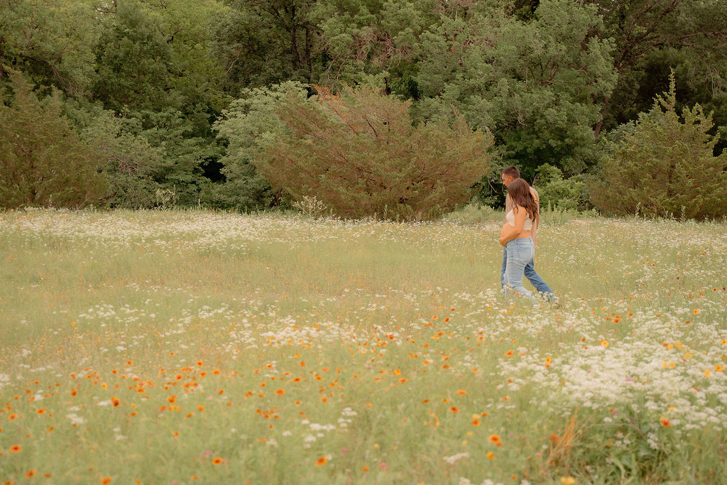 Couple walking across wildflower field during summer for their fun maternity photoshoot