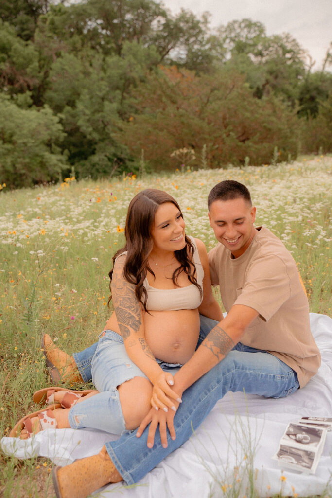 Pregnant couple sitting in a blanket in a wildflower field smiling at each other