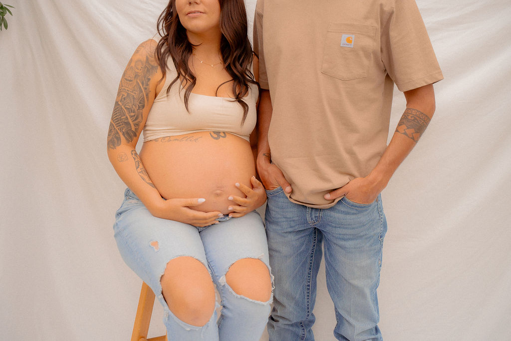 Mom holding her pregnant belly while sitting on a wooden stool and dad standing next to her