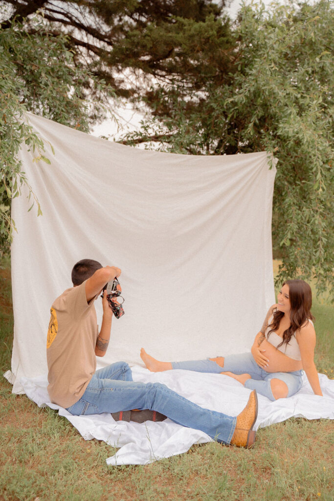 Husband taking a photos of pregnant mom on a vintage film camera under a tree